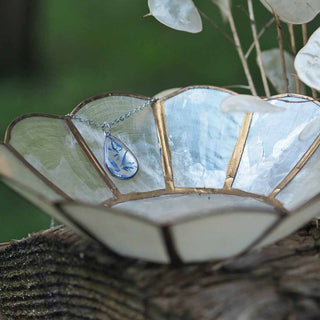 Collier en acier inoxydable argenté avec inclusion de fleurs de bleuet posé sur une coupelle blanche et dorée - collier fleur